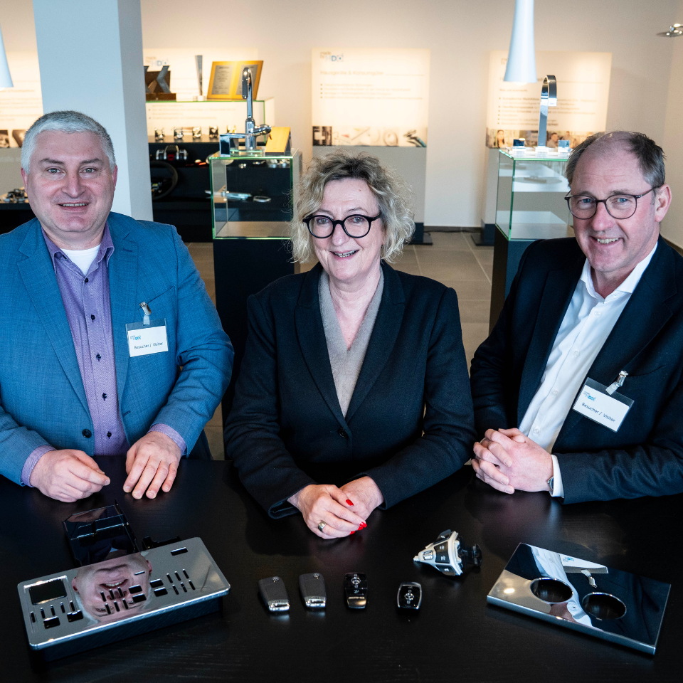 Drei lächelnde Personen mit Besucherausweisen sitzen an einem Tisch mit verschiedenen Autoteilen und elektronischen Schlüsseln in einem Ausstellungsraum.