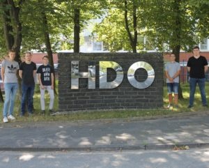 Sechs junge Männer stehen verteilt rechts und links neben einer großen Backsteinmauer mit spiegelnden, silbernen Buchstaben, die ‚HDO‘ formen, im Freien vor Bäumen und einer Straße.