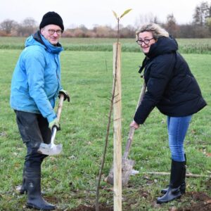 Ein Mann und eine Frau graben mit Spaten neben einem neu gepflanzten jungen Baum auf einer grünen Wiese, beide tragen Outdoor-Kleidung und Gummistiefel, im Hintergrund Bäume und ein bewölkter Himmel.