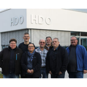 Sieben Personen stehen vor einem Gebäude mit der Aufschrift "HDO", lächeln teilweise und sind lässig gekleidet, im Hintergrund Fenster mit geschlossenen Jalousien.