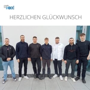 Sieben junge Männer stehen in einer Reihe vor einer weißen Wand mit der Aufschrift "Herzlichen Glückwunsch" darüber, alle leger gekleidet und in die Kamera lächelnd.