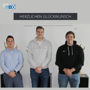 Drei junge Männer stehen nebeneinander vor einer weißen Wand mit der Aufschrift "HERZLICHEN GLÜCKWUNSCH" und dem Zitat "Permanente Weiterentwicklung ist unsere Unternehmensmaxime." Sie tragen Hemd, Sweatshirt und Hoodie und blicken freundlich in die Kamera.