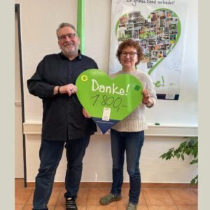 Ein Mann und eine Frau stehen vor einer Wand mit einem Poster in Herzform, sie halten zusammen ein großes grünes Herz-Schild mit der Aufschrift Danke! 1.800,-