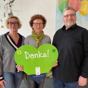 Drei Personen, zwei Frauen und ein Mann, die eine grüne herzförmige Tafel mit der Aufschrift "Danke!" halten, im Hintergrund bunte Luftballons auf einem Gemälde an der Wand