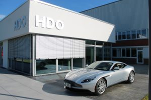 Silberner Sportwagen vor modernem weißen Gebäude mit großem HDO-Logo an der Fassade und an der Wand, das sich in der Motorhaube des Autos spiegelt