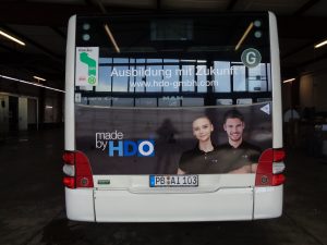 Rückansicht eines weißen Busses in einer Halle mit einer Werbebotschaft, die zwei lächelnde Menschen in schwarzen Poloshirts zeigt, auf denen das Logo "HDO" zu sehen ist, dazu der Schriftzug "made by HDO" und der Hinweis "Ausbildung mit Zukunft www.hdo-gmbh.com"