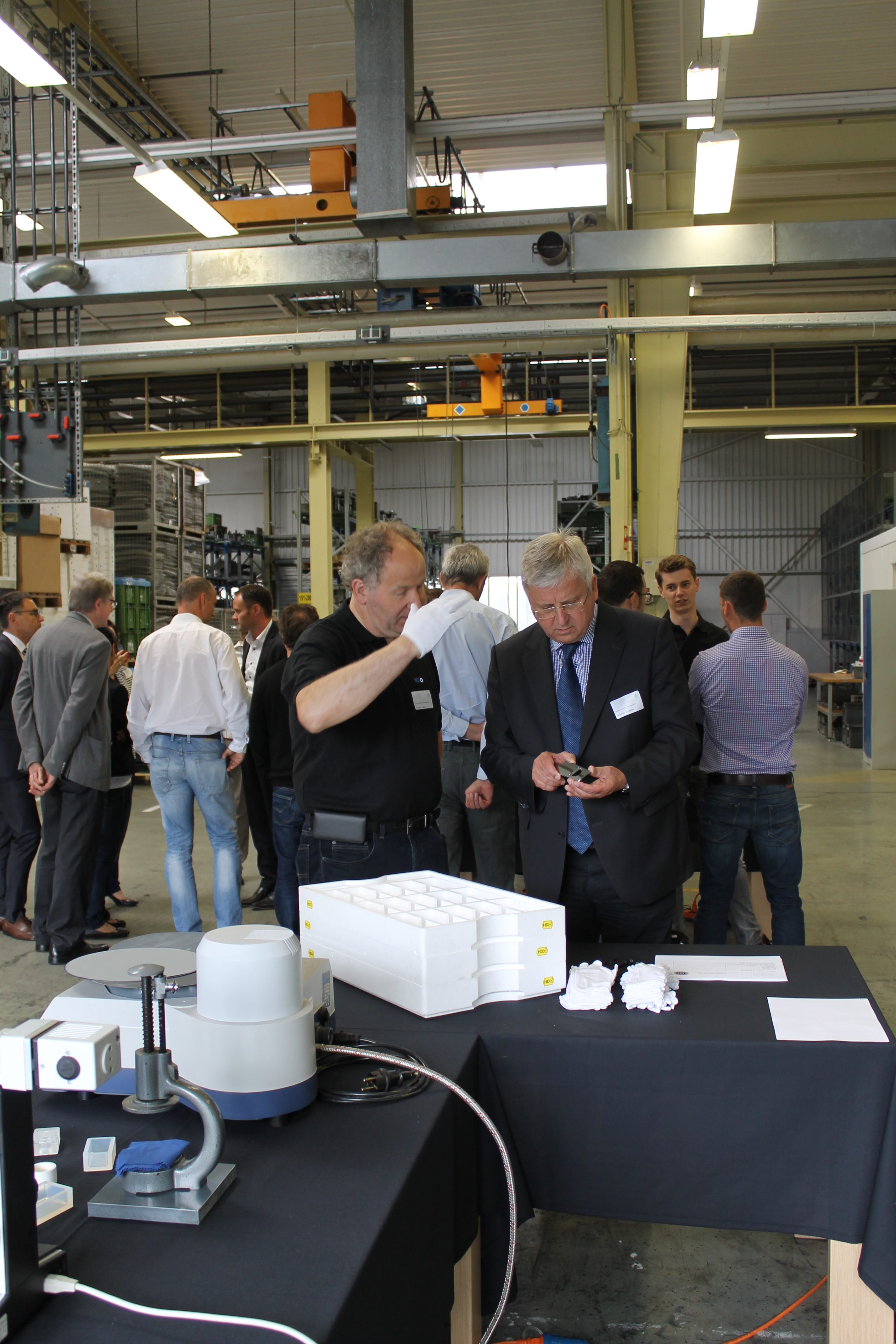 Menschen in einer Fabrikumgebung, zwei Männer schreiten ein technisches Gerät gemeinsam durch und tragen Namensschilder mit der Aufschrift "HDO", im Vordergrund ein Tisch mit technischen Geräten und weißen Handschuhen mit "HDO"-Markierungen