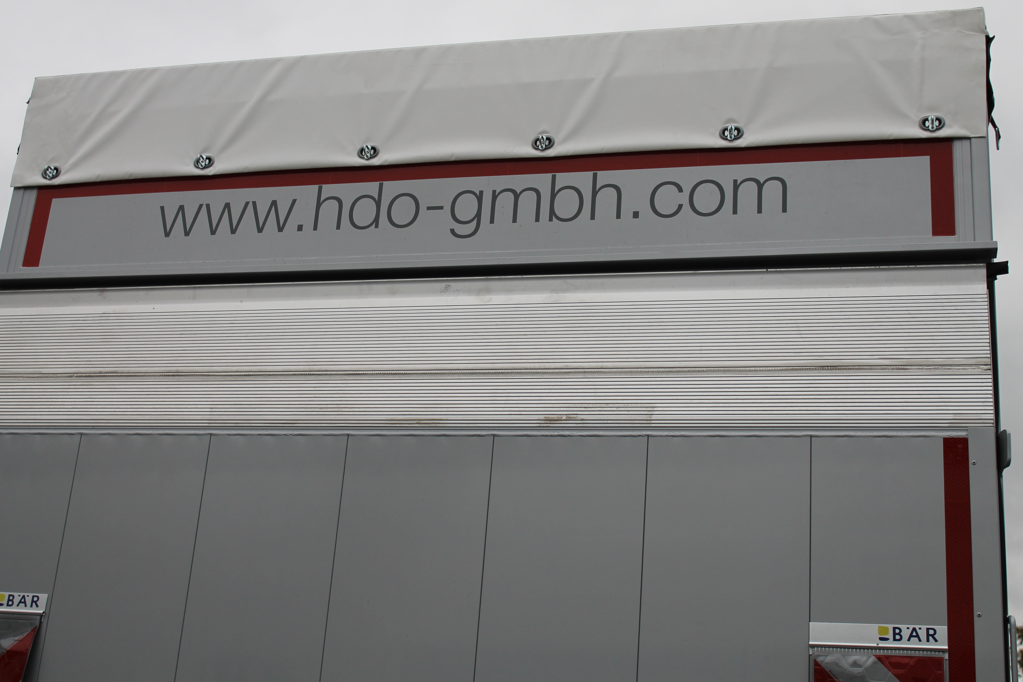 Rückseite eines Lastwagens mit der Webseite www.hdo-gmbh.com als Aufschrift am oberen Rand der Ladefläche und einem weißen Planenverdeck mit Befestigungselementen oben.