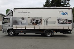 Lieferwagen mit Werbebanner, das verschiedene metallische Produkte zeigt, darunter einen Seifenspender, ein Fahrpedal, einen Wasserhahn und einen Autoscheinwerfer, mit dem Schriftzug "made by HDO" und der Webseite www.hdo-gmbh.com