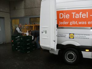 Zwei Männer laden grüne Kisten aus einem Transporter mit geöffneten Türen an einer Gebäudewand, auf dem Transporter ist ein oranger Schriftzug "Die Tafel – Jeder gibt, was er kann" zu sehen.