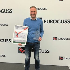 Mann in blauem Pullover und Jeans steht vor einem weißen Hintergrund mit dem Schriftzug EUROGUSS, hält eine gerahmte Urkunde und eine gläserne Auszeichnung in den Händen und lächelt in die Kamera.