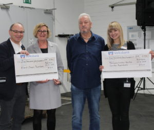 Vier Personen stehen nebeneinander, zwei von ihnen halten große Schecks der Volksbank über jeweils 2500 Euro, mit der Aufschrift HDO, Klinik-Clowns Paderborn und Therapeutisches Reiten Caritas Paderborn; linker Mann mit Brille und dunklem Sakko, daneben Frau mit hellgrauem Mantel, dritter Mann in dunkelblauer Hemd und Jeans, rechte Frau mit blonden Haaren und schwarz-weiß gestreiftem Oberteil, HDO als Spender auf den Schecks erkennbar