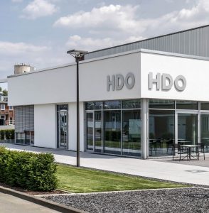 Modernes weißes Gewerbegebäude mit großen Glasscheiben und dem Schriftzug "HDO" an zwei Seiten der Fassade, umgeben von gepflegtem Grün und einem Weg mit laternenbeleuchtetem Gehweg unter bewölktem Himmel.