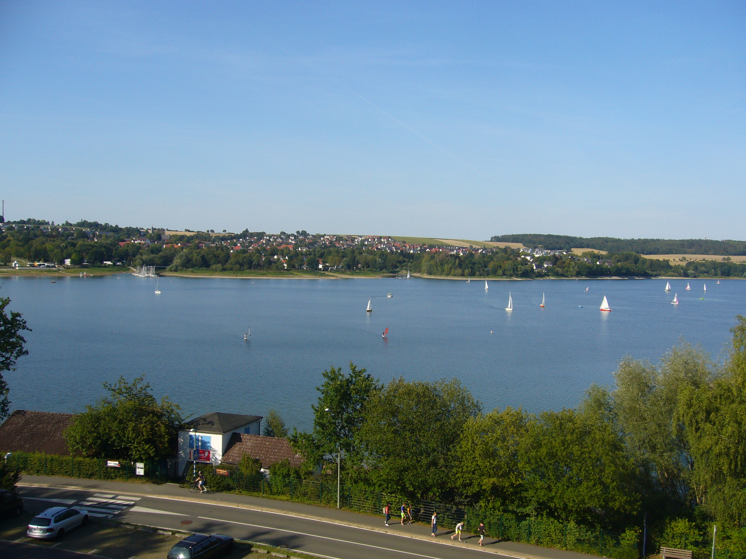 Panoramablick auf einen großen See mit zahlreichen Segelbooten bei klarem Himmel, im Vordergrund Bäume, eine Straße mit Fußgängern und Autos sowie ein Gebäude mit sichtbarem 