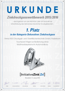Urkunde für den Zinkdruckgusswettbewerb 2015/2016 mit dem 1. Platz in der Kategorie Dekorativer Zinkdruckguss für die Firma HDO Druckguß- und Oberflächentechnik GmbH, Paderborn, für einen Touchpad-Träger zur Bedienung eines Multimediasystems im Automobilbau, inklusive Abbildung des preisgekrönten Zinkdruckguss-Teils