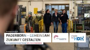 Drei Personen, zwei Frauen und ein Mann, gehen gemeinsam durch eine Werkstatt oder Fabrikhalle, im Vordergrund unscharf eine weitere Person, dazu Schrift mit dem Slogan Paderborn - Gemeinsam Zukunft gestalten und Logos der Wirtschaftsförderung Paderborn und HDO.